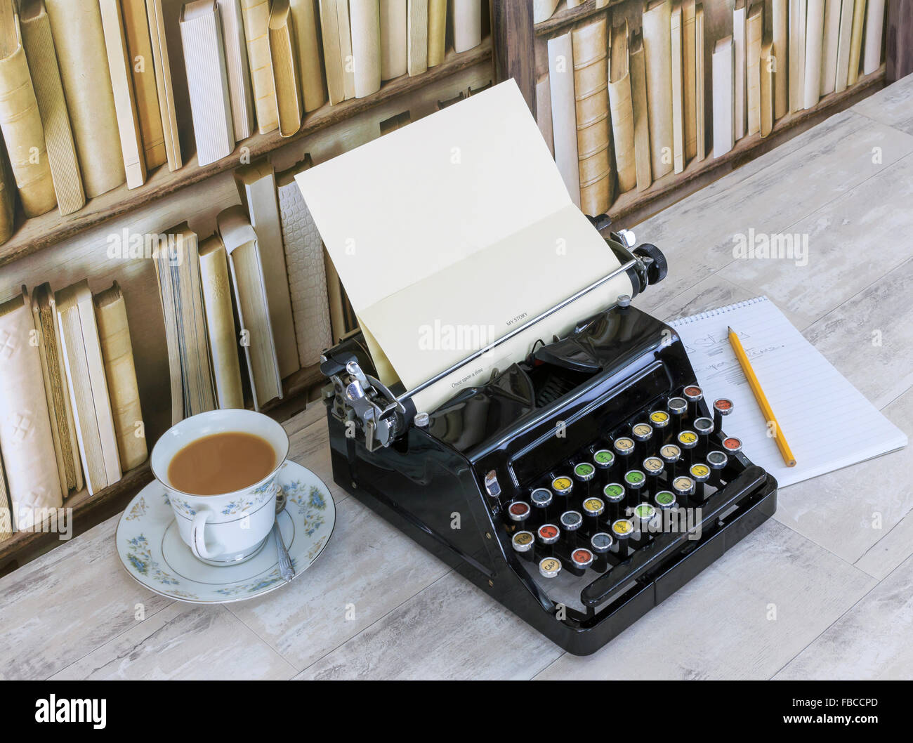 Vintage typewriter, notepad, pencil and china teacup with tea on old shabby wood effect desktop with old bookcase background Stock Photo