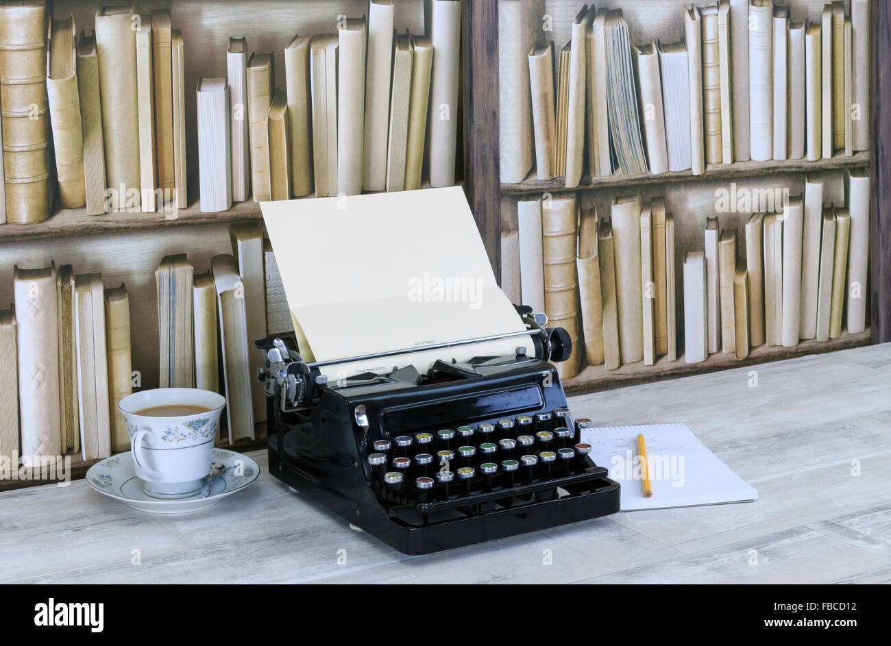Vintage typewriter, notepad, pencil and china teacup with tea on old shabby wood effect desktop with old bookcase background Stock Photo