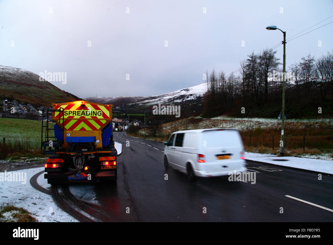 Nant y Moel, Mid Glamorgan, South Wales. 15th January 2016: A van on the A4061 road passes a gritting truck as it enters the village of Nant y Moel in the upper Ogmore Valley in South Wales, with with fresh snowfall on the Mynydd Llangeinwyr hills behind. Several cm of snow fell on the hills overnight, prompting the Met Office to issue a yellow level severe weather warning of snow and ice for the region. Stock Photo