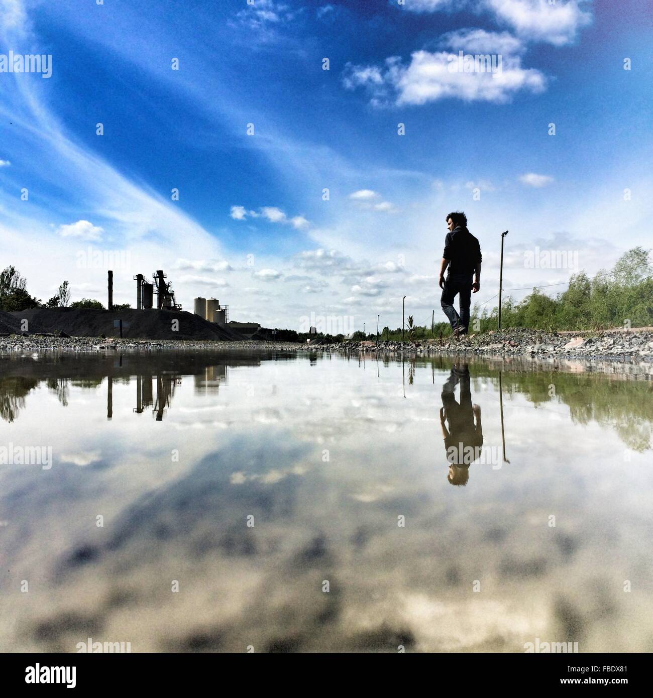 Reflection Of Young Man Walking At Riverbank Against Blue Sky Stock Photo