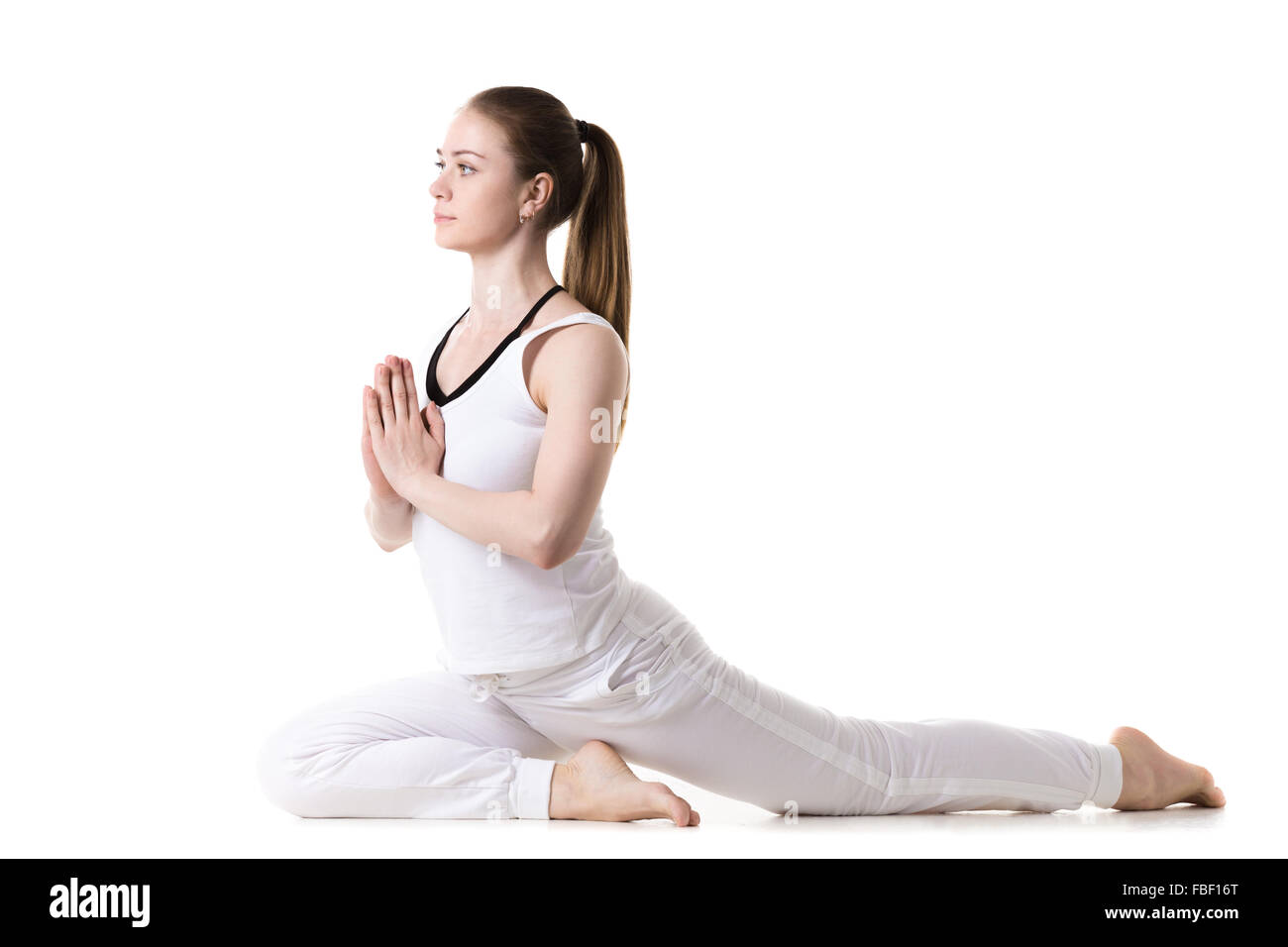Young fitness model in white sportswear doing yoga or pilates training, sitting in Half pigeon pose (ardha kapotasana), side vie Stock Photo