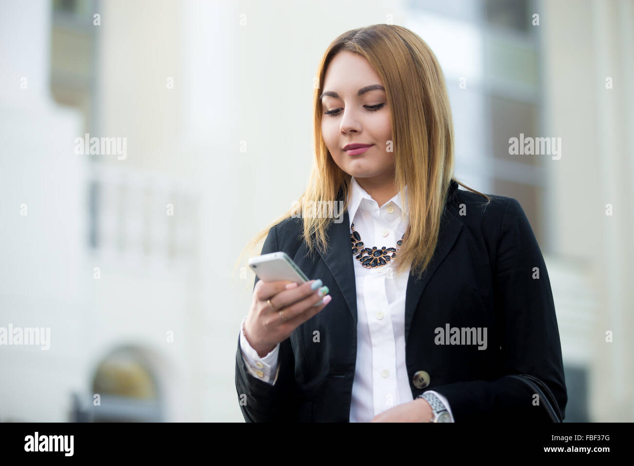 Beautiful young woman wearing formal business suit, walking on city street, holding smartphone, making a call, using app, copy s Stock Photo