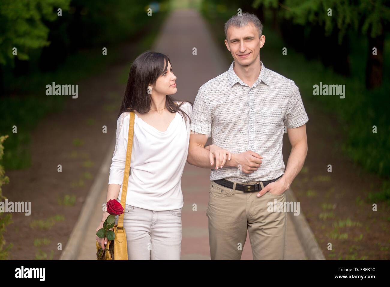 Portrait of couple in love walking together on a date on summer park walkway, young woman with yellow handbag looking at her boy Stock Photo