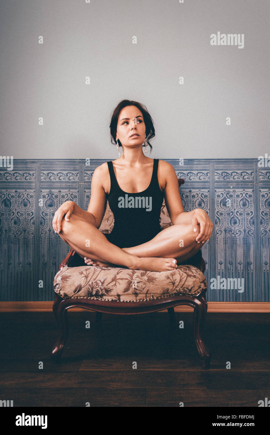 Young Woman With Legs Crossed Sitting On Armchair Against Wall At Home Stock Photo