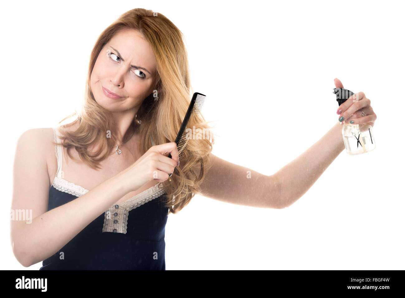 Funny frowning young attractive blond beautician woman holding spray bottle, hairdresser scissors and comb, unhappy with her hai Stock Photo