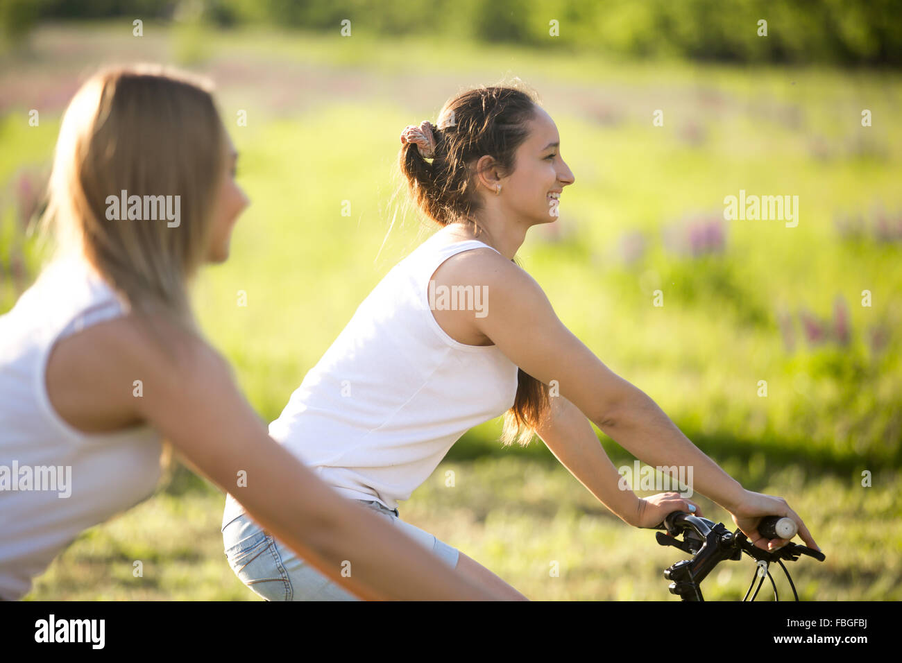 Two cute young happy smiling beautiful girlfriends wearing casual white tank tops enjoy cycling in park on sunny summer day, foc Stock Photo