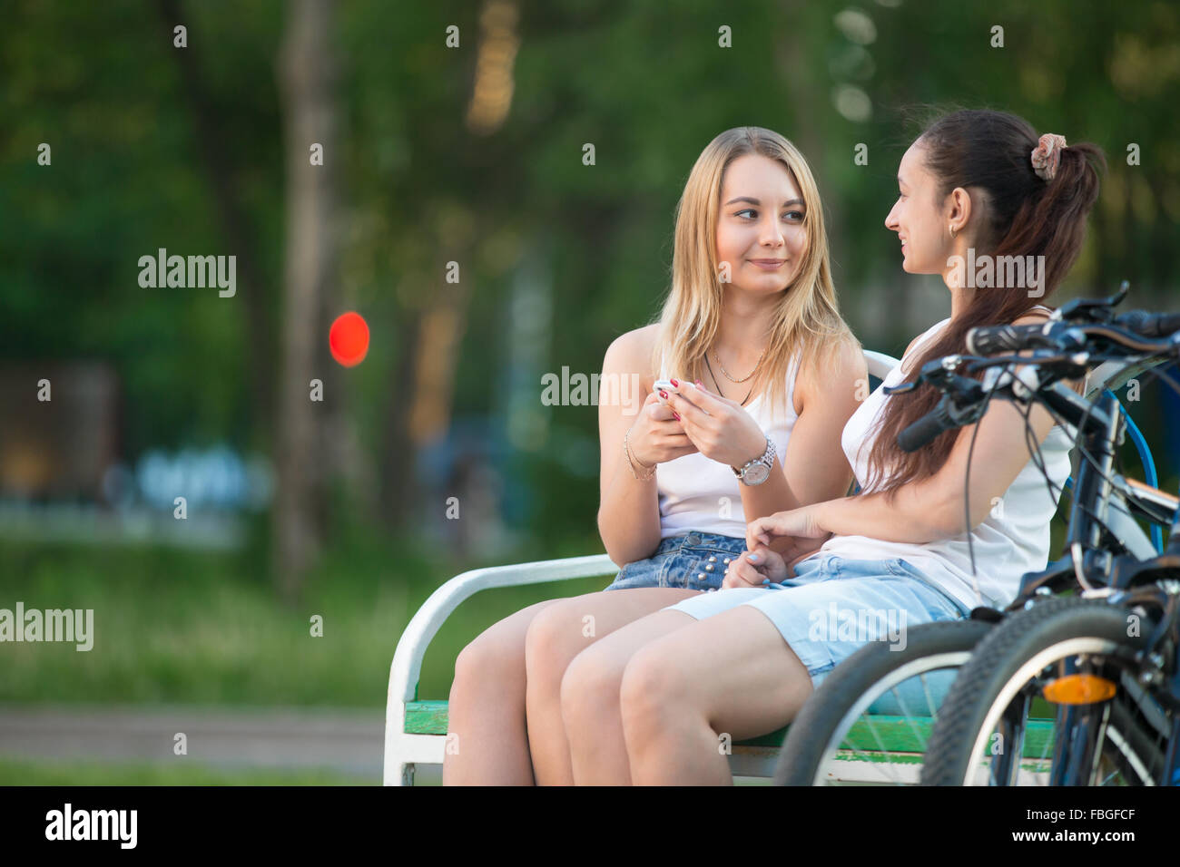 Two happy smiling beautiful young biker women friends wearing casual clothes sitting on park bench on summer day, chatting, havi Stock Photo