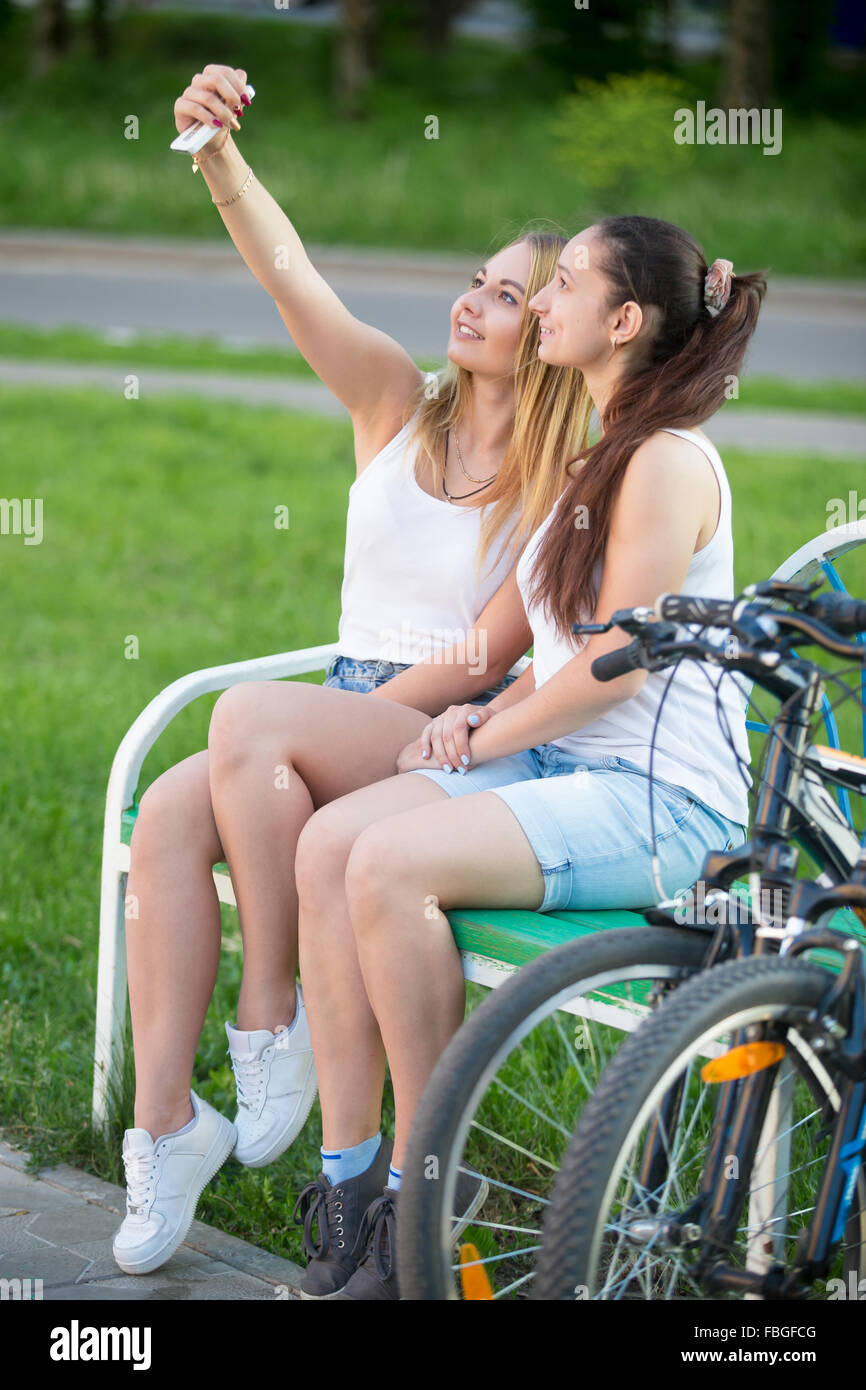 Profile view of two cute smiling beautiful bicyclist teenage girls wearing casual clothes sitting on park bench on summer day, p Stock Photo