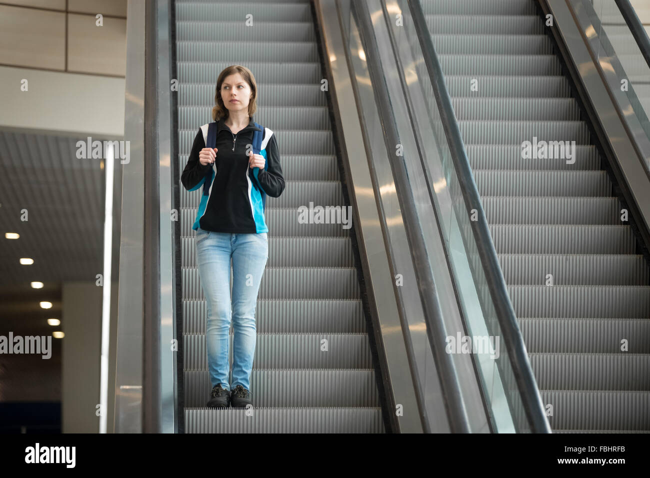 Young woman in 20s with backpack riding escalator in airport terminal, travelling alone, wearing casual style clothes Stock Photo