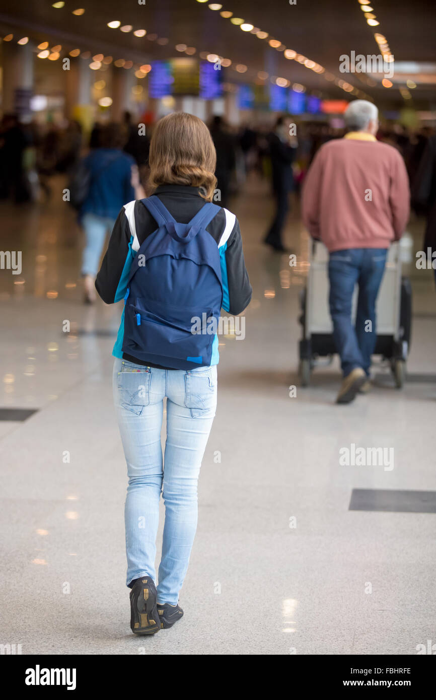 Young woman in 20s with backpack walking in airport terminal, wearing jersey, jeans and sneakers, blurred crowd of travelling pe Stock Photo