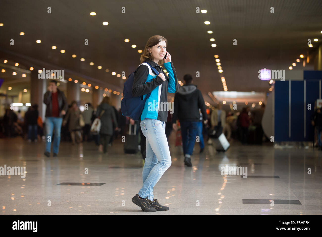 Smiling young woman in 20s with backpack walking in airport terminal, using cell phone, making call, wearing jersey, jeans and s Stock Photo