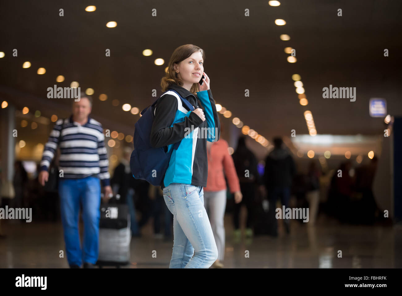 Portrait of smiling young woman in 20s with backpack walking in airport terminal, using cell phone, making call, wearing jersey Stock Photo