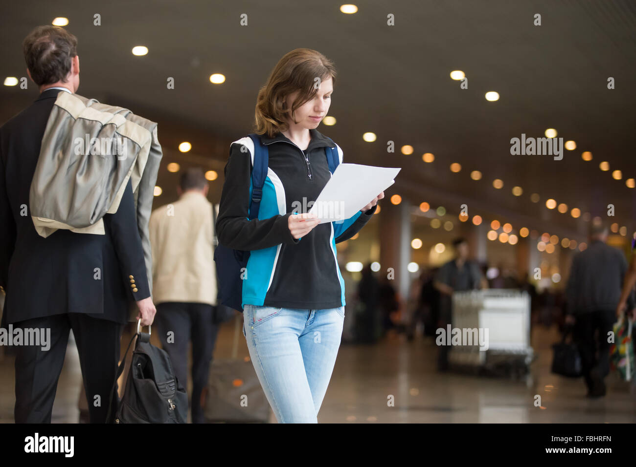 Portrait of young woman in 20s with backpack standing in airport terminal, with printed e-tickets, using cell phone to check in, Stock Photo