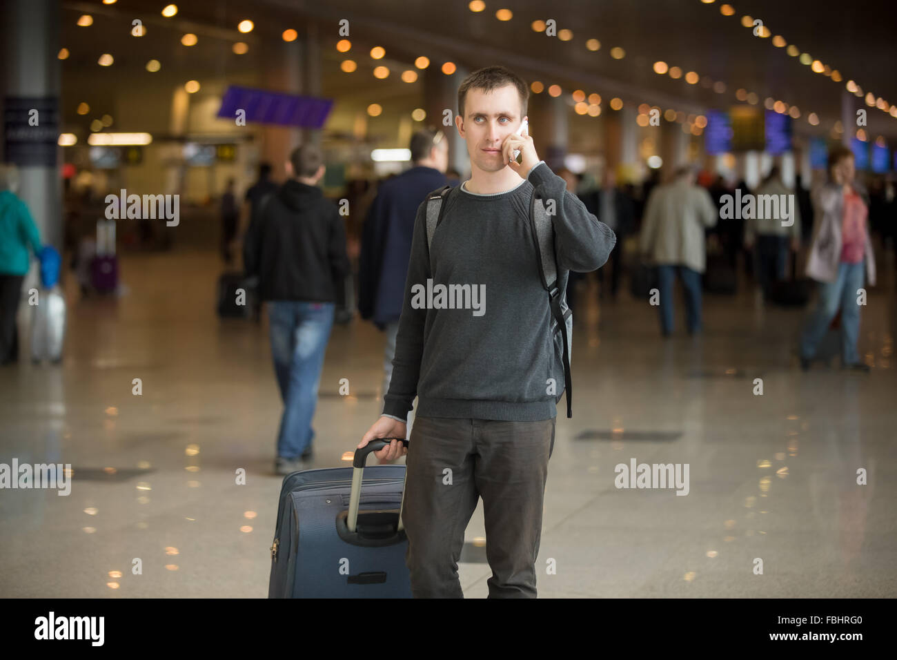 Portrait of young handsome man in 20s walking in modern airport terminal, talking on smart phone, travelling with luggage bag, w Stock Photo