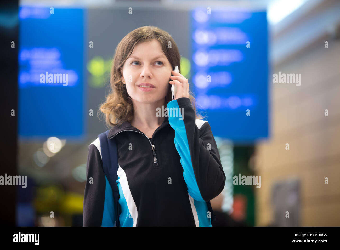 Portrait of smiling young woman in 20s with backpack walking in airport terminal, using smart phone, talking on mobile phone, bl Stock Photo