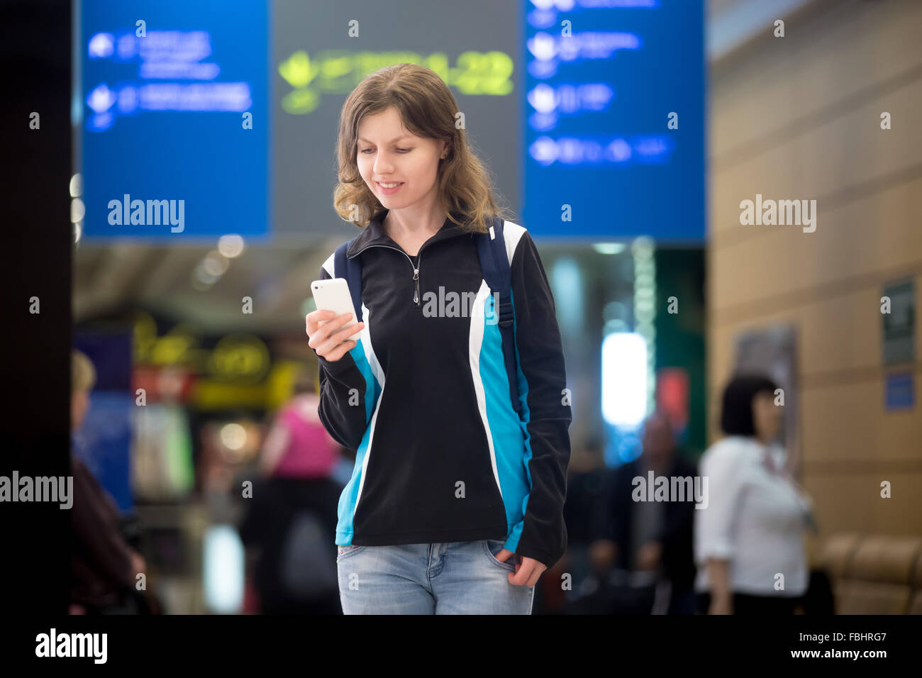 Portrait of smiling young woman in 20s with backpack waiting for flight in modern airport terminal, using cell phone app in publ Stock Photo
