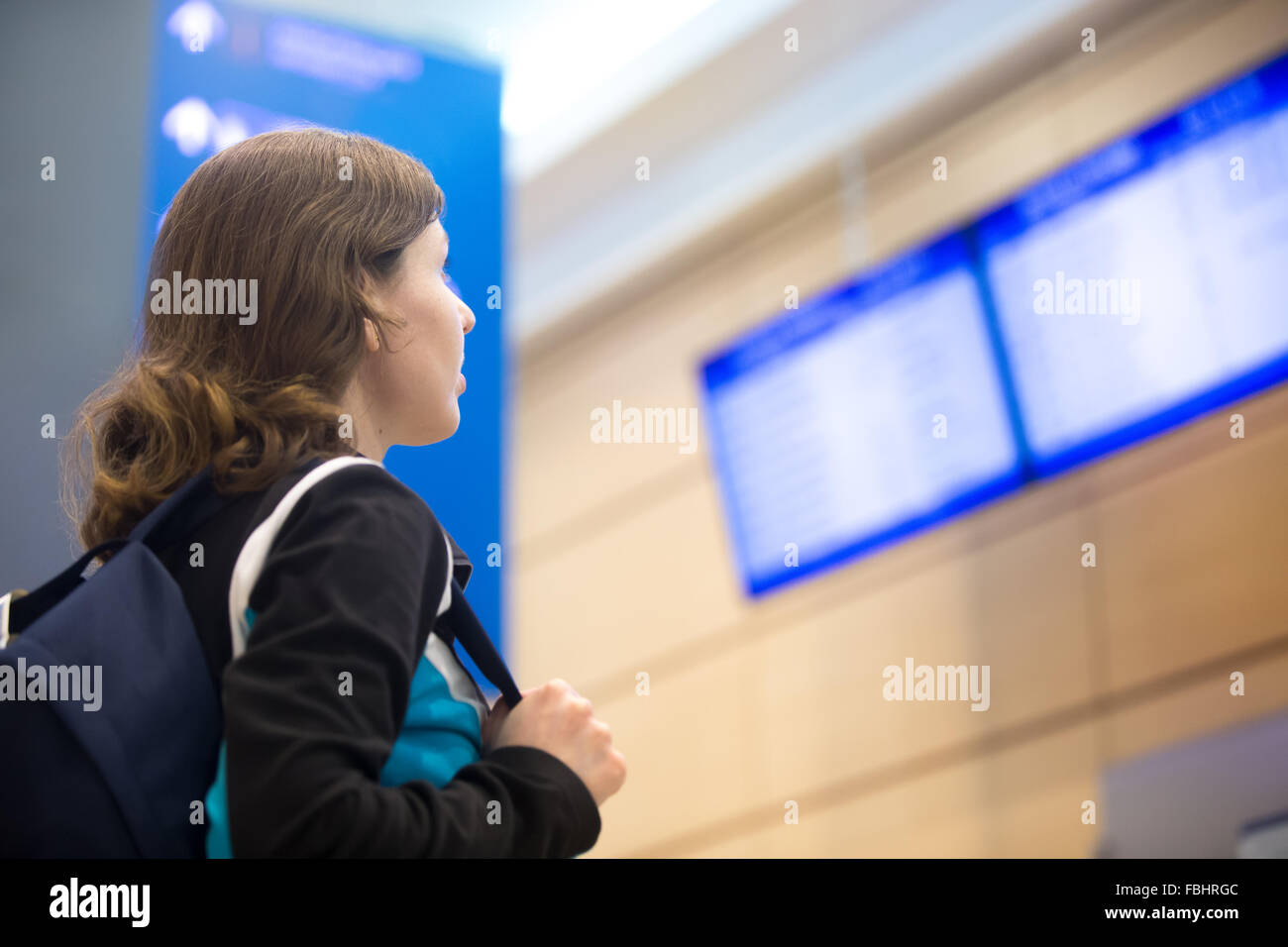 Young woman in 20s with backpack waiting for flight in modern airport terminal, looking at flight information board Stock Photo