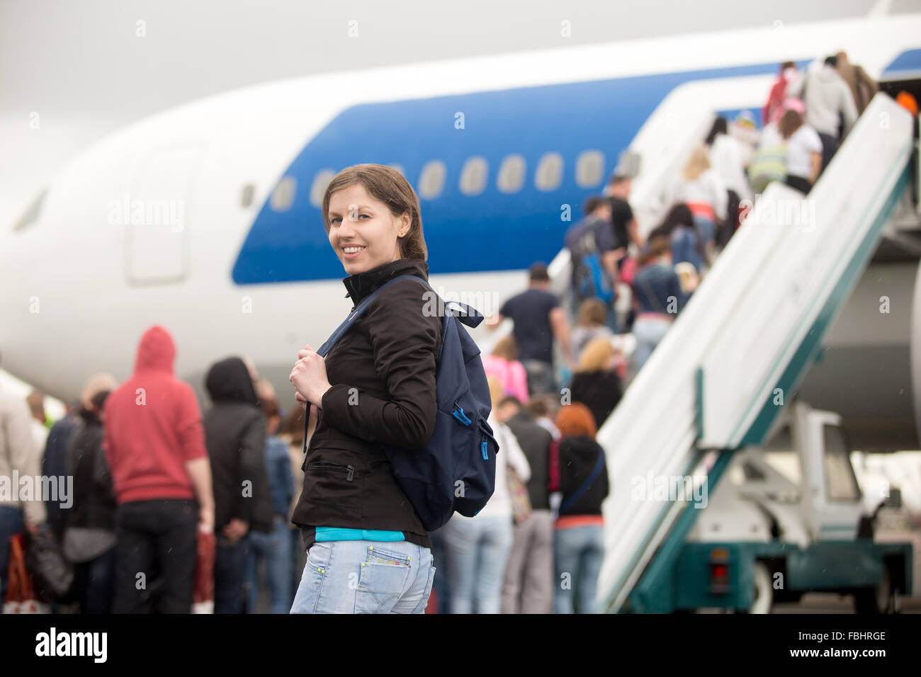 Young Caucasian happy smiling woman passenger in 20s travelling with backpack, boarding airplane, looking at camera, people clim Stock Photo