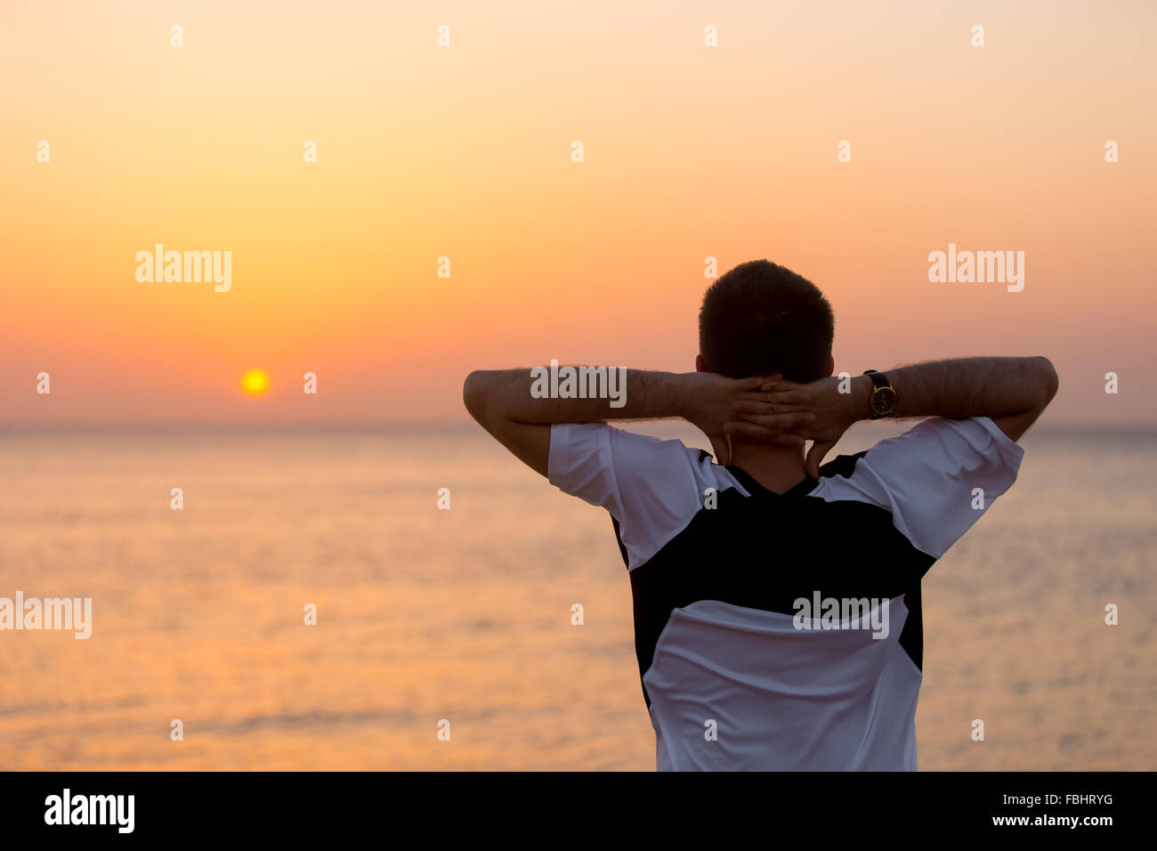 Young man standing at the seashore against colorful sunrise or sunset sky in carefree pose with hands behind his head, enjoying Stock Photo