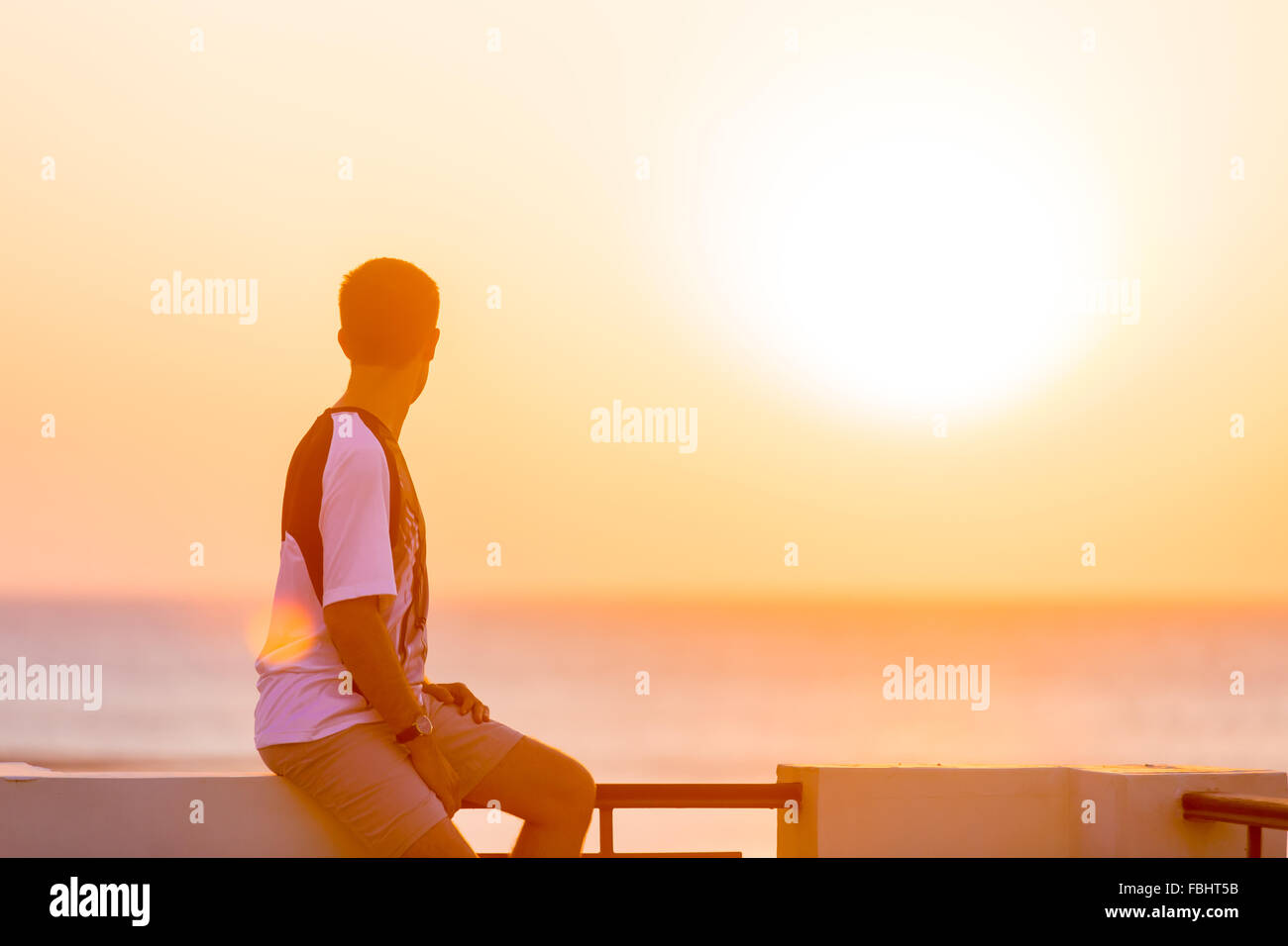 Young handsome man sitting on the balcony rail in carefree pose, enjoying beautiful colorful sunrise or sunset sea scenery and f Stock Photo