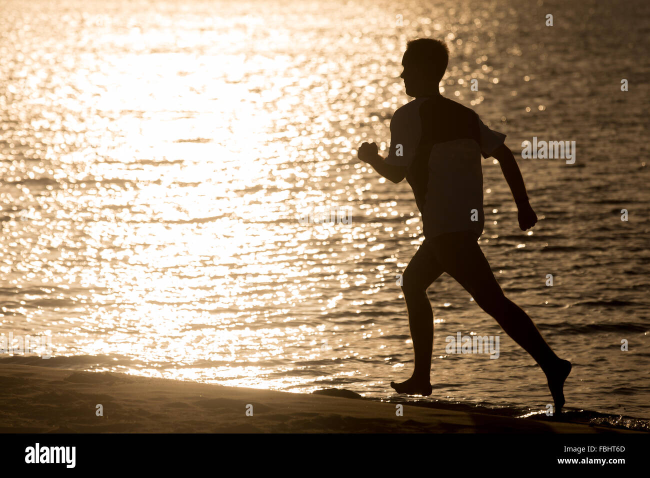 Healthy lifestyle: silhouette of sporty young man working out on the beach, running fast on the sand shore against the sea light Stock Photo