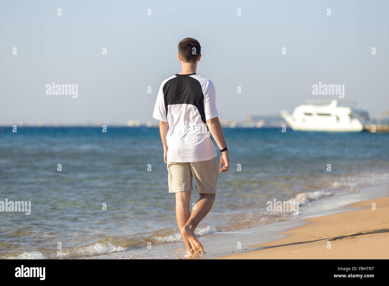 Young Caucasian sporty man walking on the coastline, making splashes, looking at the sea, white yacht on the background, back vi Stock Photo