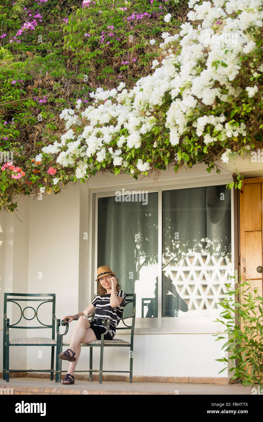 Young woman in cute straw hat and summer dress sitting on chair on terrace of white cottage house with flower roof, using cell p Stock Photo