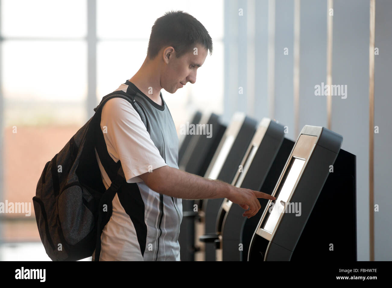 Young man with backpack touching interactive display at self-service transfer machine, doing self-check-in for flight or buying Stock Photo