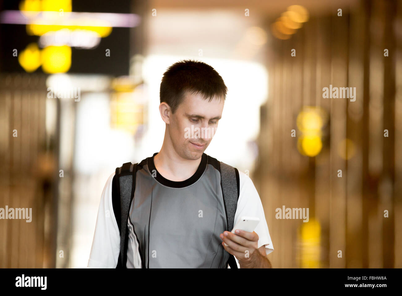 Smiling young man with backpack in his 20s walking in modern building, holding smartphone, looking at screen, using cell phone a Stock Photo
