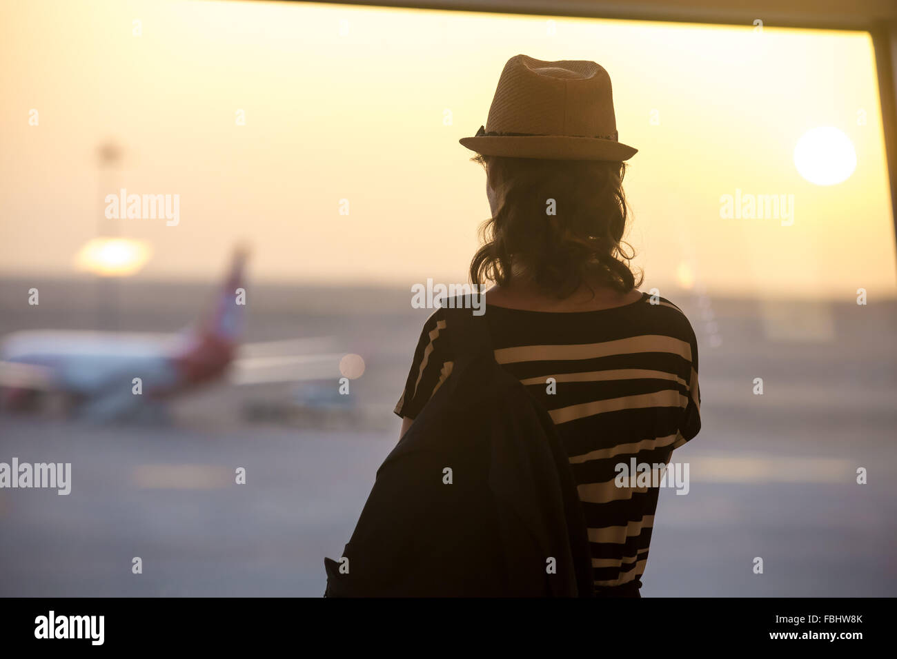 Young woman in straw hat and summer dress with backpack waiting for flight in modern airport terminal building, watching airplan Stock Photo
