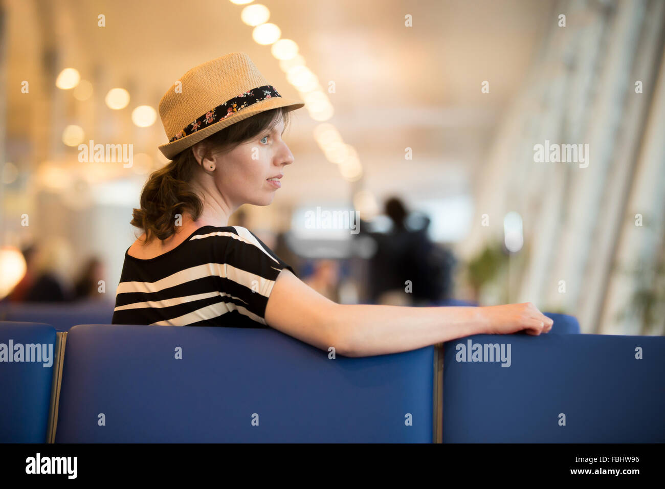 Profile portrait of young traveling woman in summer dress and straw hat waiting for trip, sitting with thoughtful expression in Stock Photo