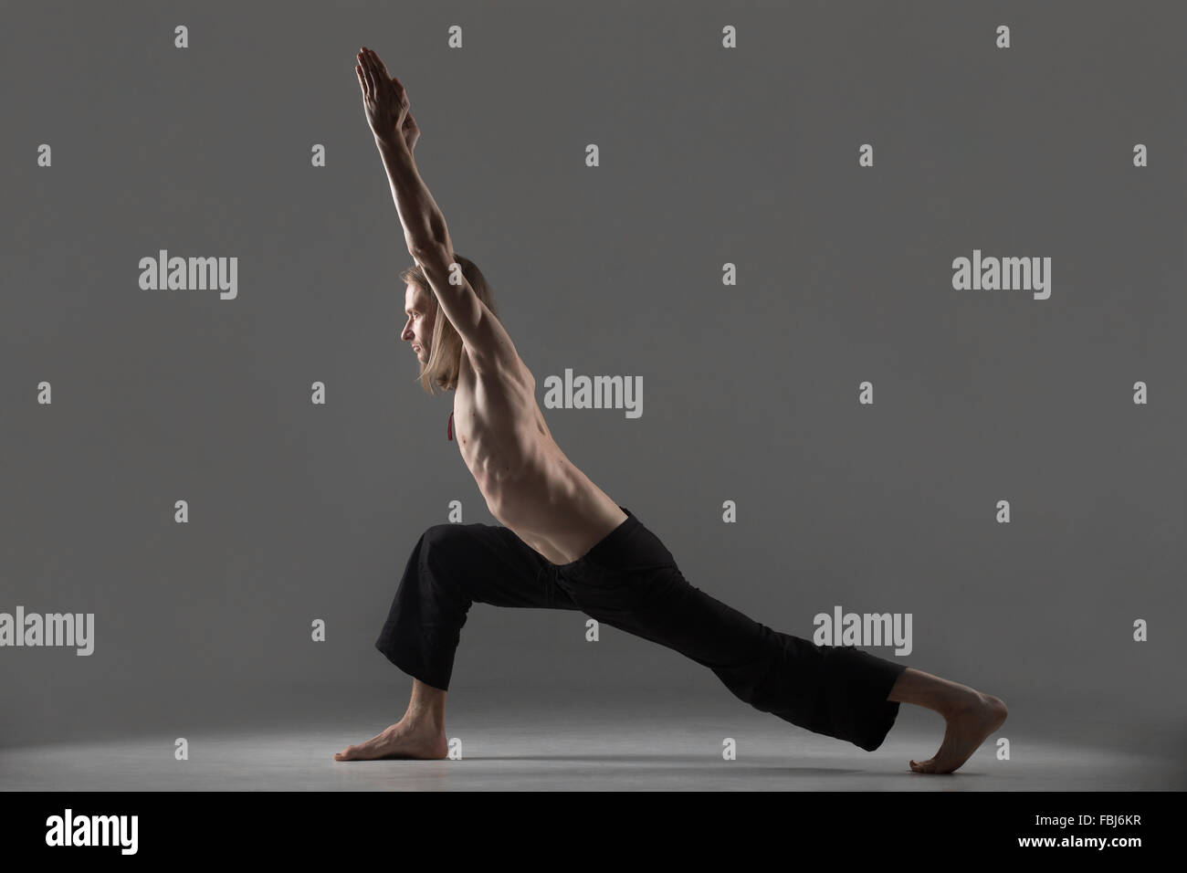 Sporty young man in black trousers doing lunge exercise, standing in variation of Warrior I posture, Virabhadrasana 1, profile v Stock Photo