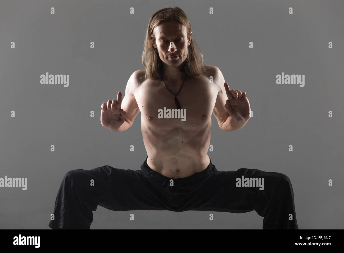 Portrait of sporty young man in black pants doing strengthening yoga pose, standing in rudrasana (sumo), utkata konasana (goddes Stock Photo