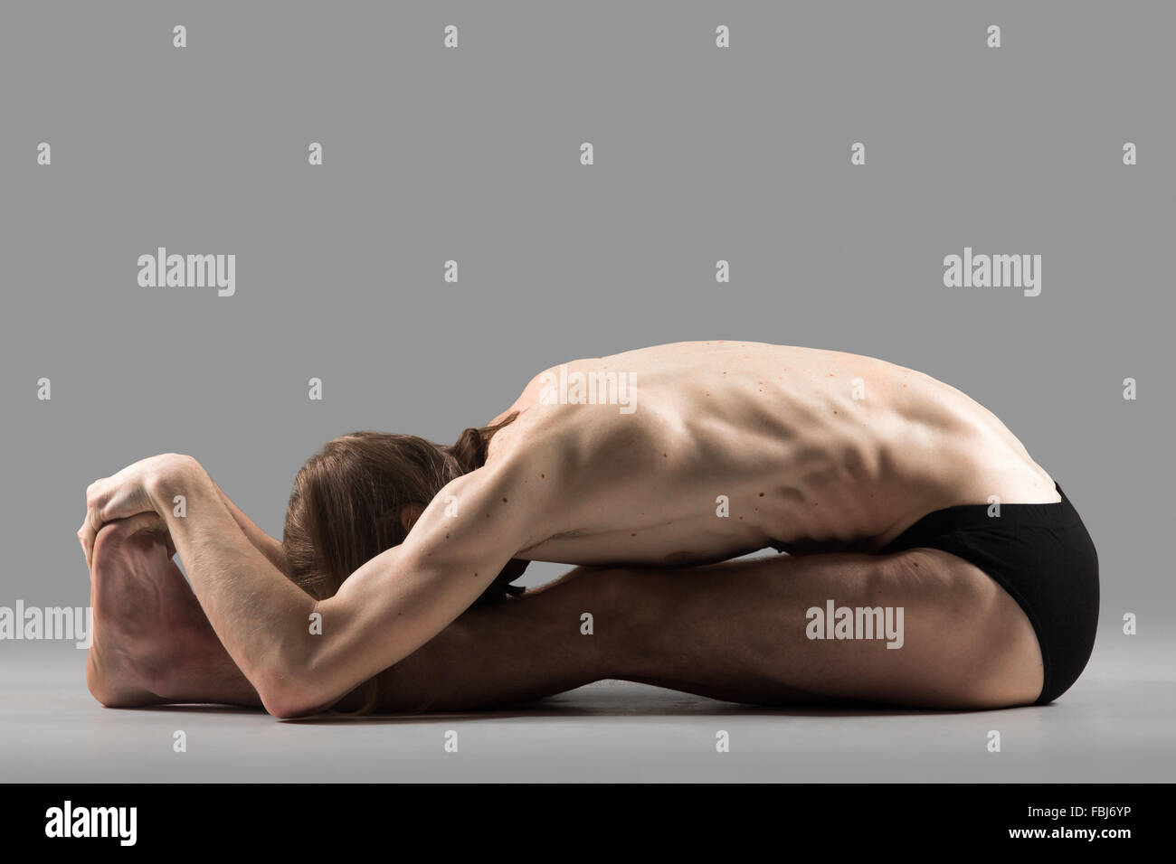 Sporty muscular young yogi man doing sit-up, sitting in paschimottanasana pose, seated forward bend posture, studio shot on dark Stock Photo