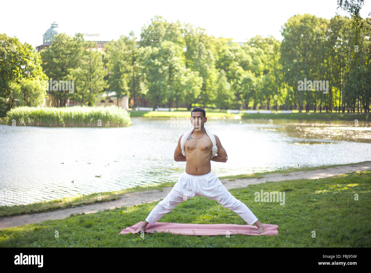 Attractive Indian young man in white linen clothes working out on river bank in park, doing preparation for Pyramid Pose, Intens Stock Photo