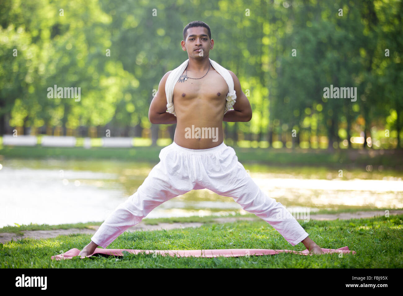 Attractive Indian young man in white linen clothes working out on riverbank in park, doing preparation for Pyramid Pose, Intense Stock Photo