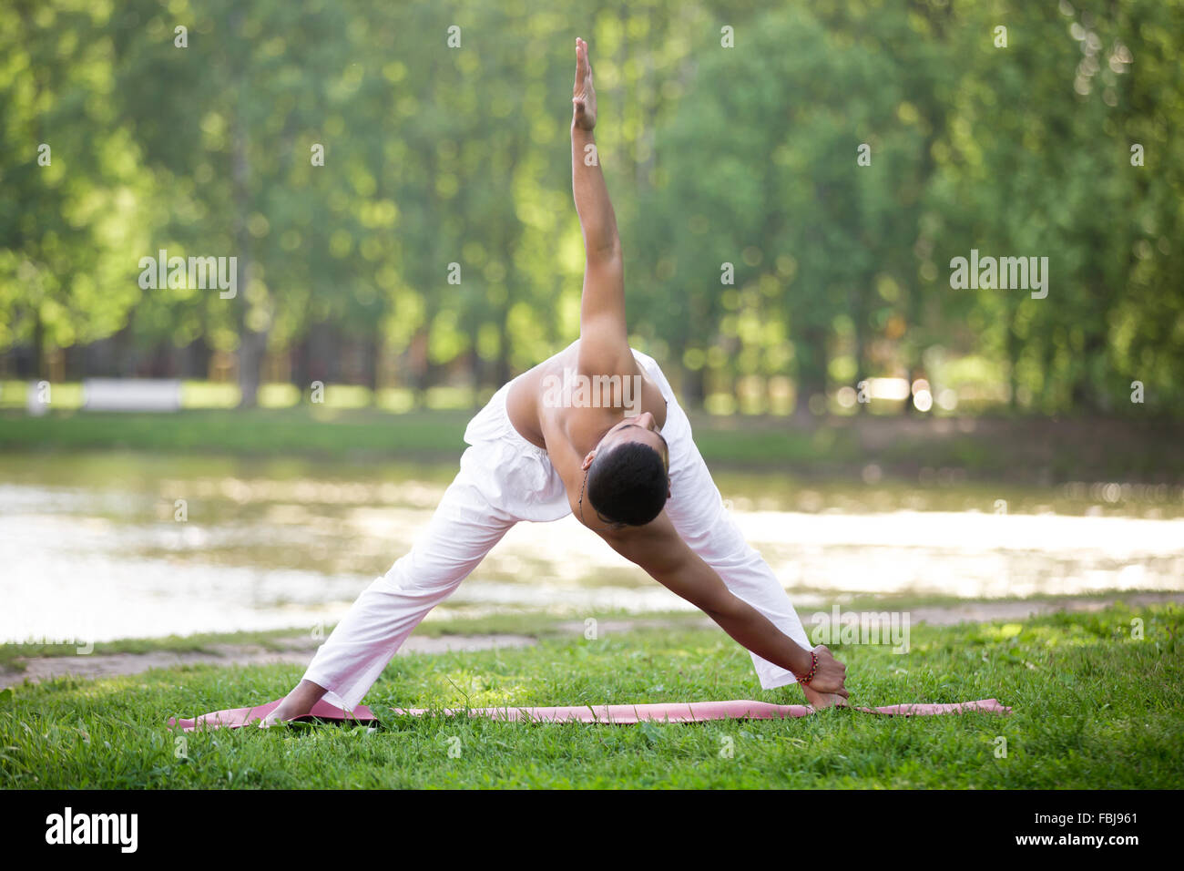 Attractive Indian young yogi man in white linen clothes working out on river bank in park, standing in Cross Triangle posture va Stock Photo
