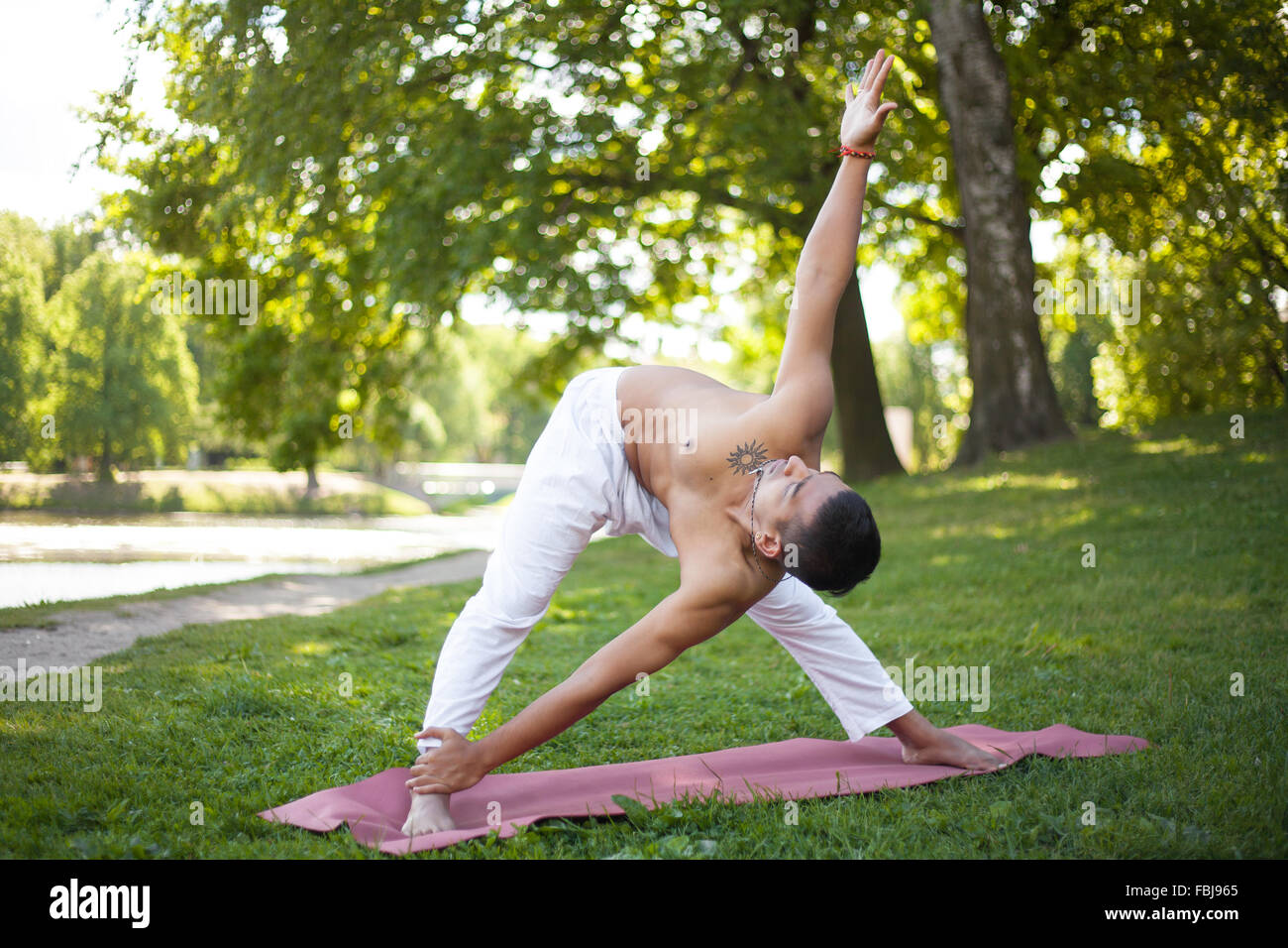 Attractive asian young yogi man in white linen clothes working out on lake in park, standing in Cross Triangle posture modificat Stock Photo