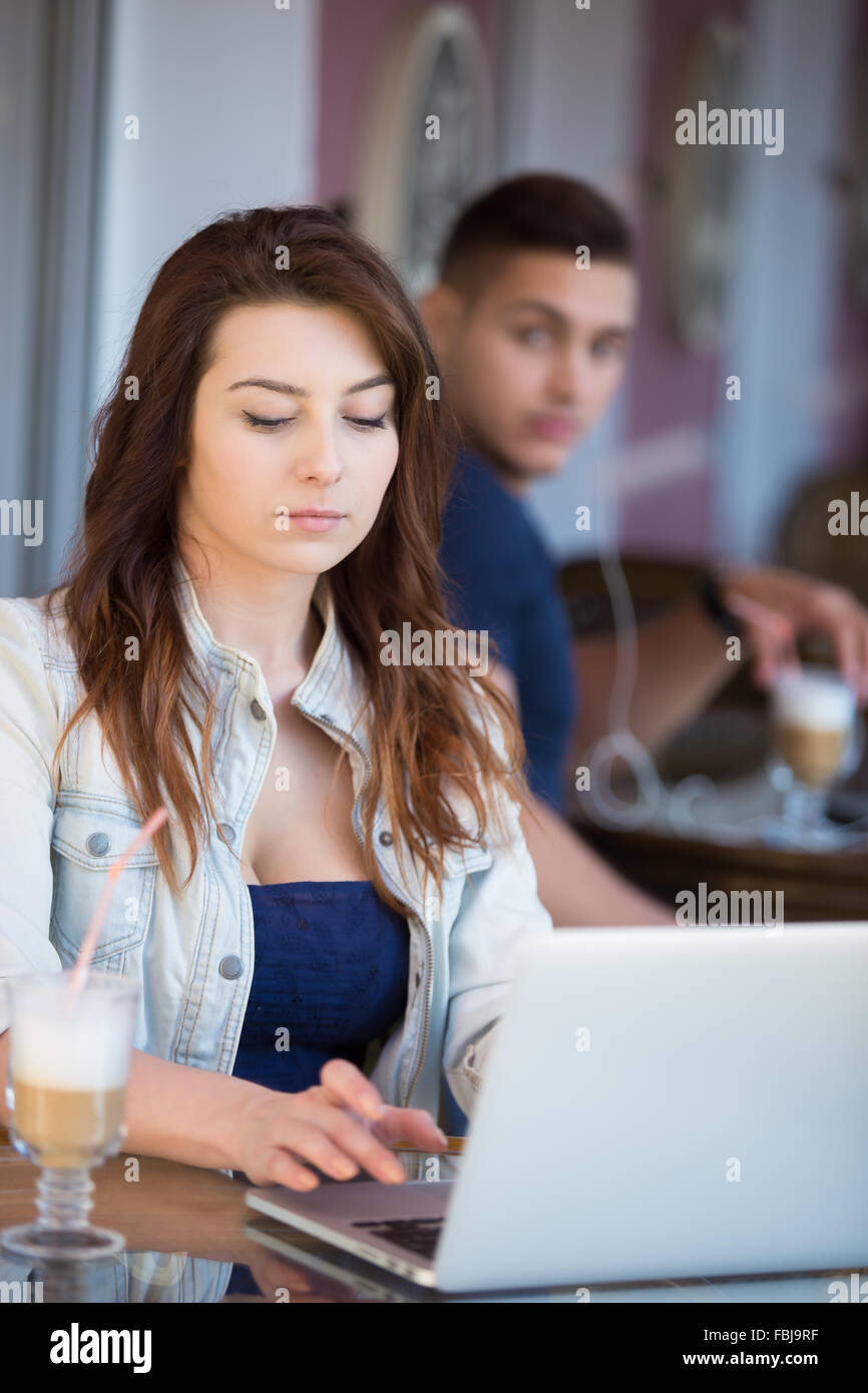 Beautiful serious young woman in casual clothes sitting in street cafe working on laptop while being watched by handsome guy beh Stock Photo