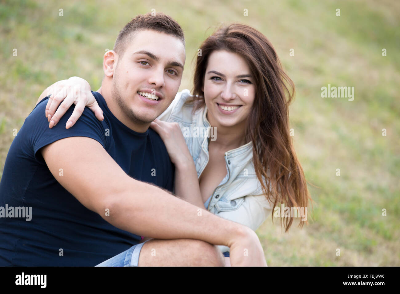 Portrait of beautiful happy smiling young couple on a date embracing, sitting together on grass in park on summer day Stock Photo