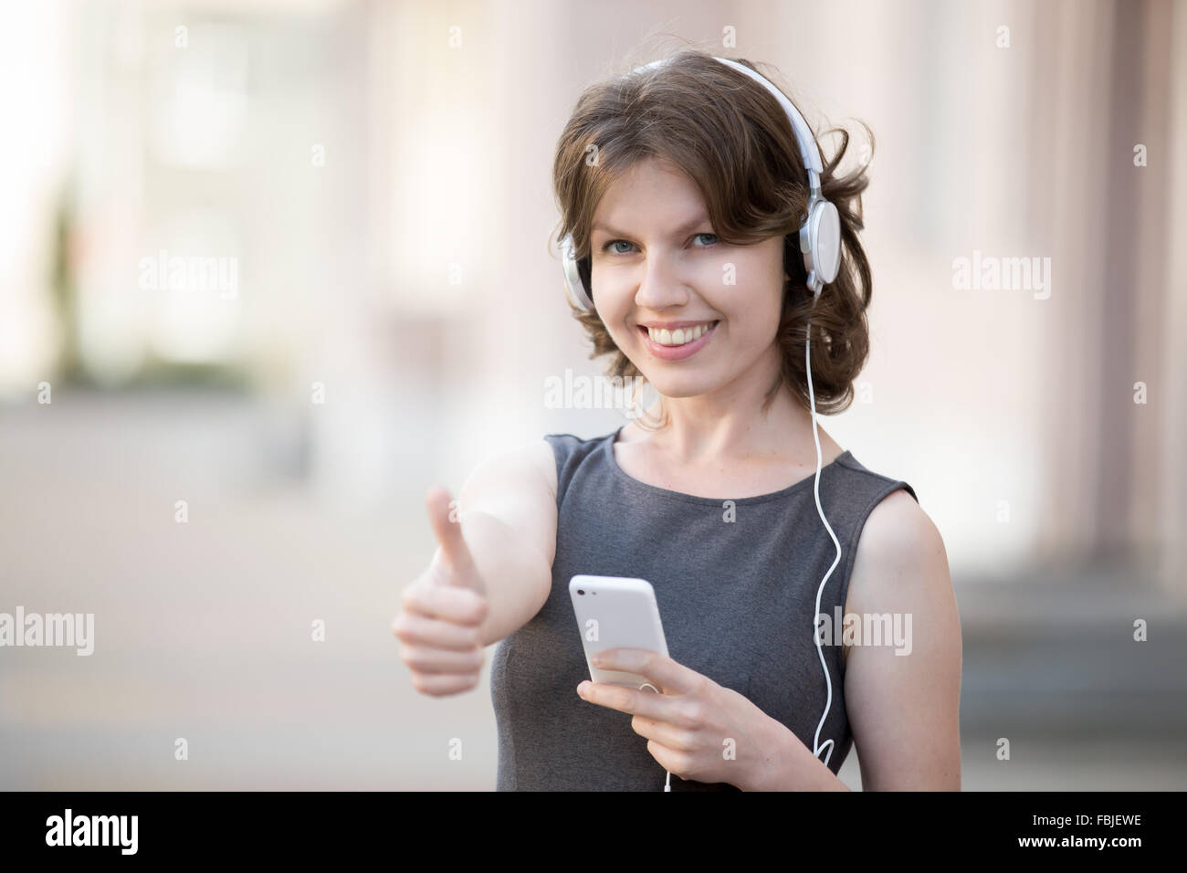 Portrait of happy woman in white earphones holding cellphone in hands on summer street, listening music, friendly smiling Stock Photo