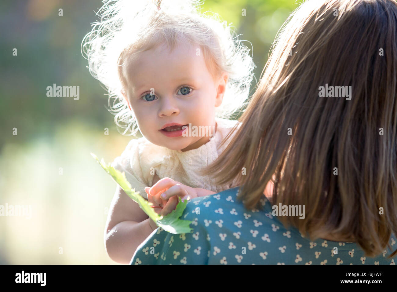 Portrait of happy adorable baby girl with her young mother playing with tree leaf, mom holding her little daughter in arms, walk Stock Photo