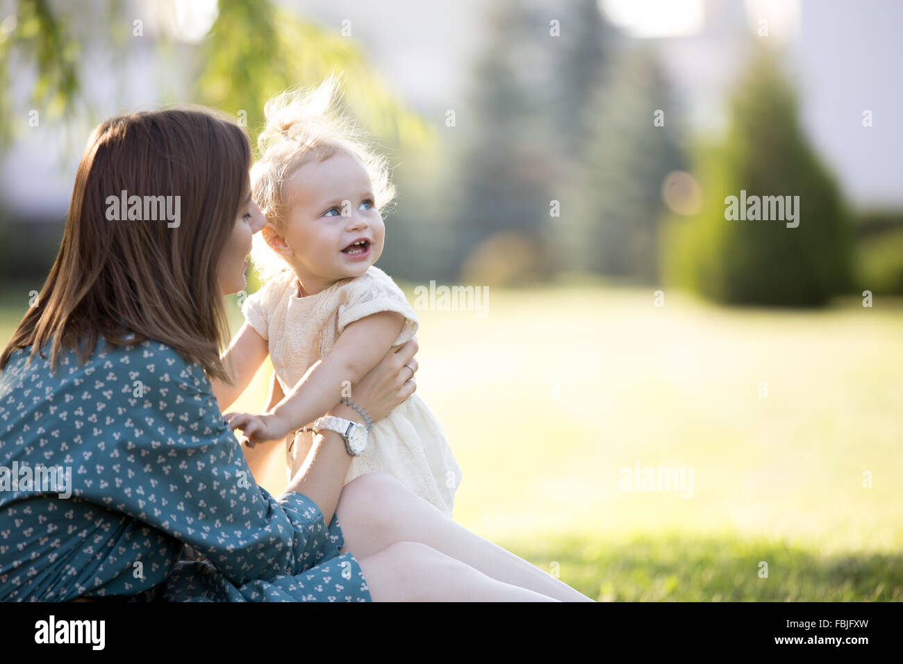 Happy young mom and adorable blond girl playing together in park in summertime, smiling mother holding her little daughter in ar Stock Photo