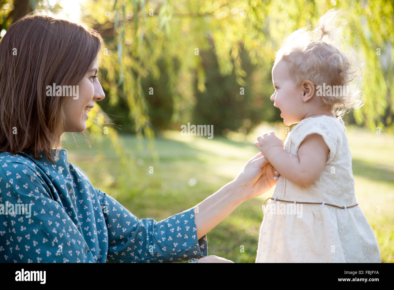 Happy young mom and adorable blond girl playing together in park in summertime, smiling mother holding her little daughter hand, Stock Photo