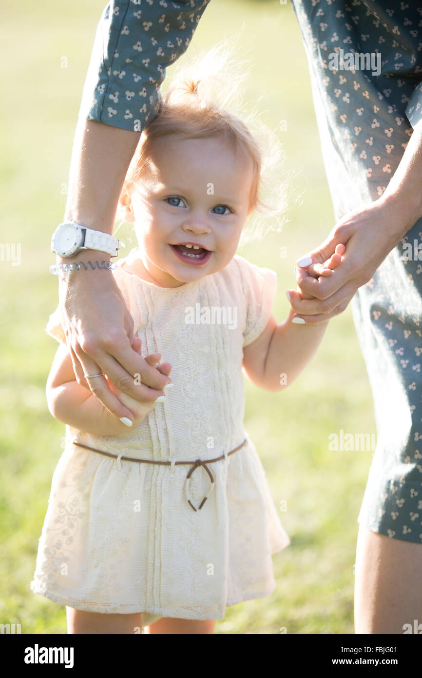 Young mom teaching to walk cheerful adorable blond toddler girl in white dress, mother holding her little daughter hands, suppor Stock Photo