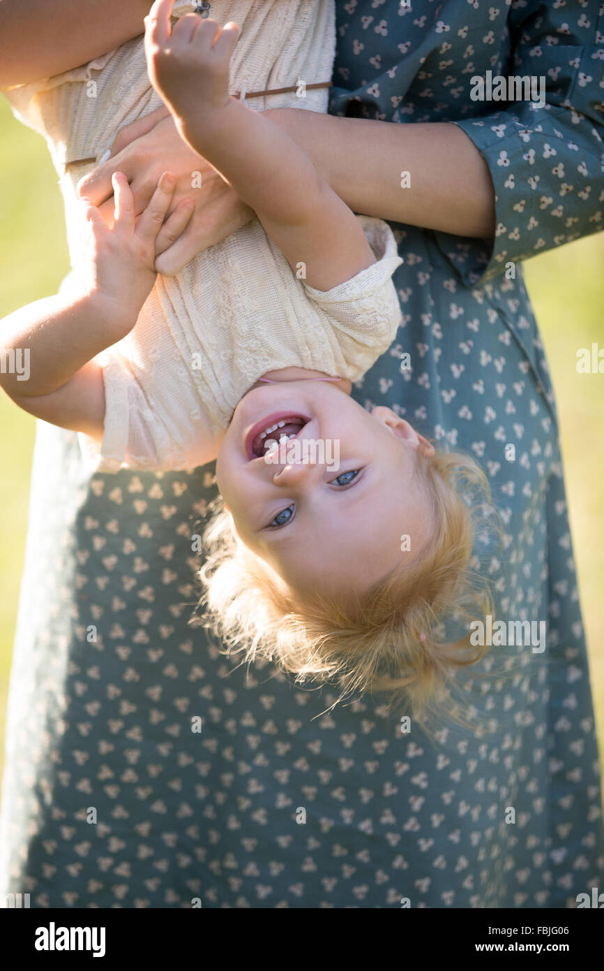 Young mom and cheerful adorable blond tot girl playing, having fun together in park in summertime, mother playfully holding her Stock Photo