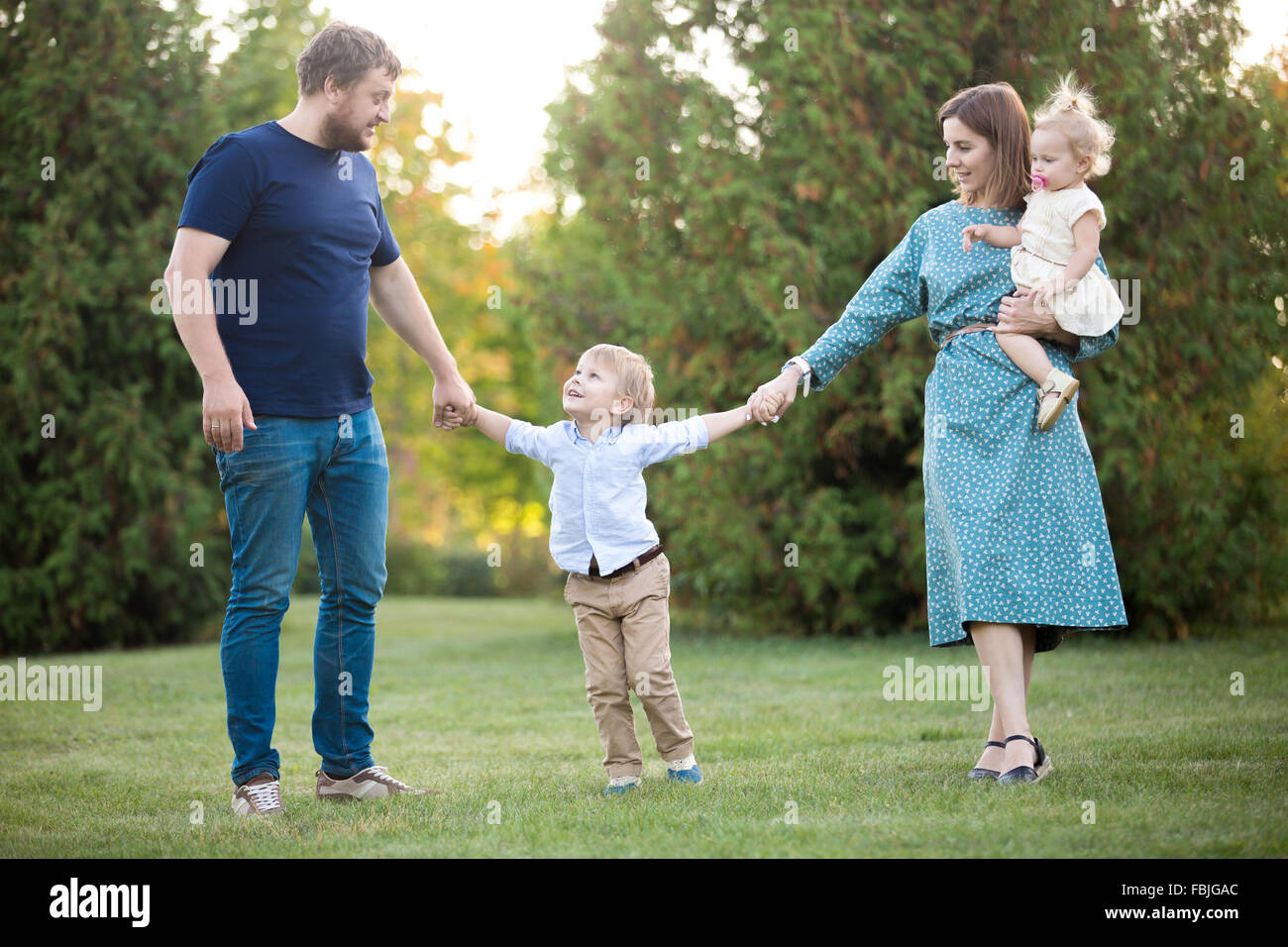 Portrait of happy beautiful family of four walking in park. Mom, dad and two siblings enjoying leisure time in summer, talking, Stock Photo