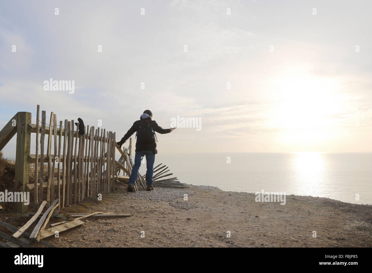 A man holds onto a broken fence whilst taking a picture over the edge the cliff at Beachy Head with sea and sun in the distance. Stock Photo