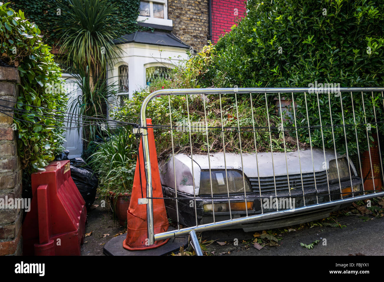 An abandoned car lays under a mountain of grass and refuge in a Dublin city front garden Stock Photo