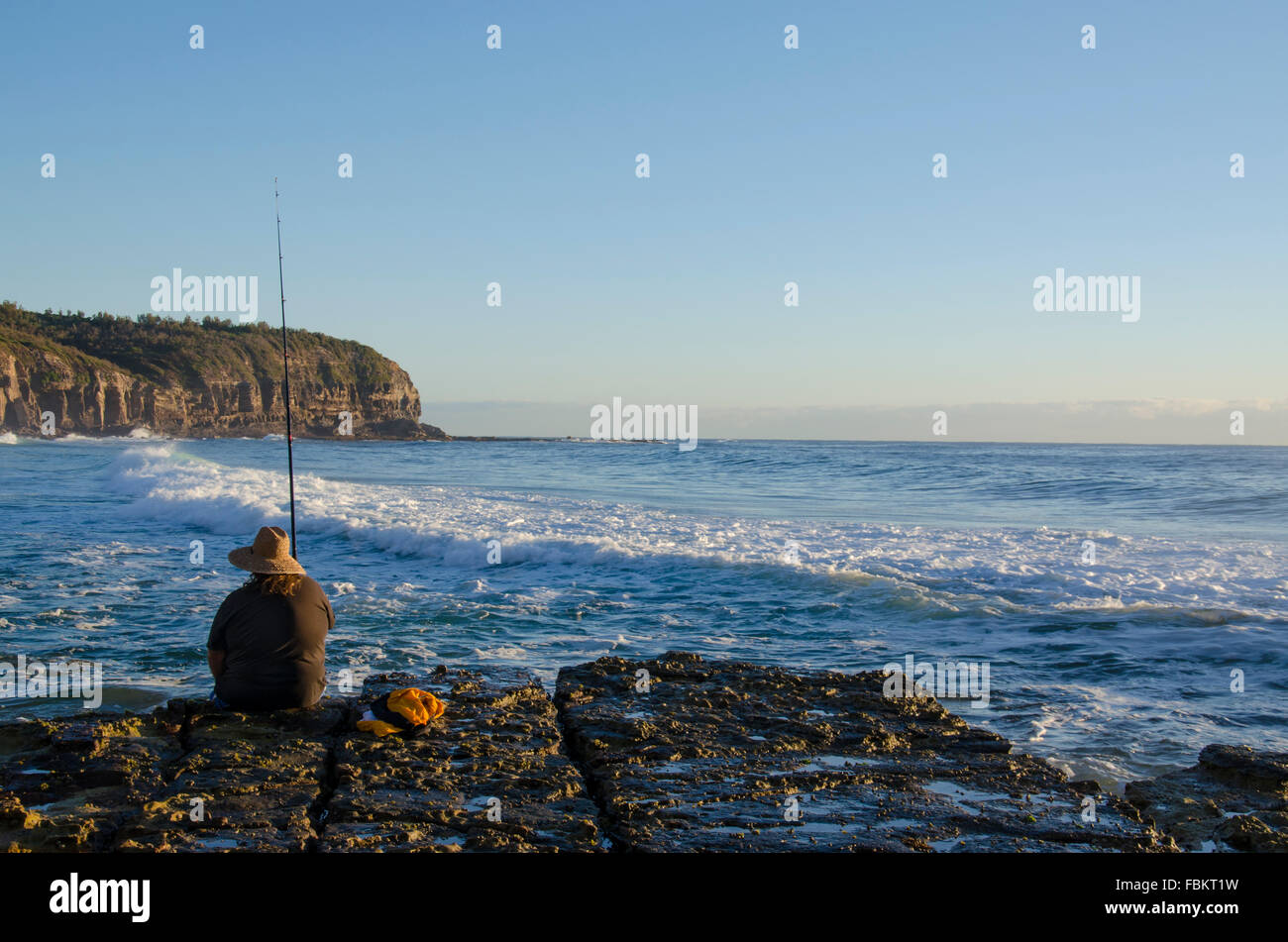 A Caucasian person fishes at dawn at the edge of a rocky platform as waves roll in and the sun rises on a Sydney beach Stock Photo